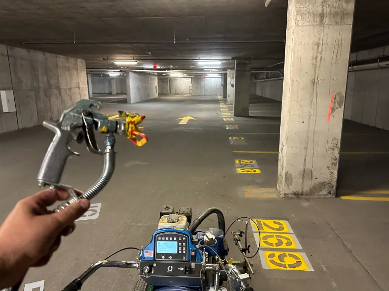 Firstperson view of operating a blue pressure washer in a dim underground parking garage with numbered parking bays