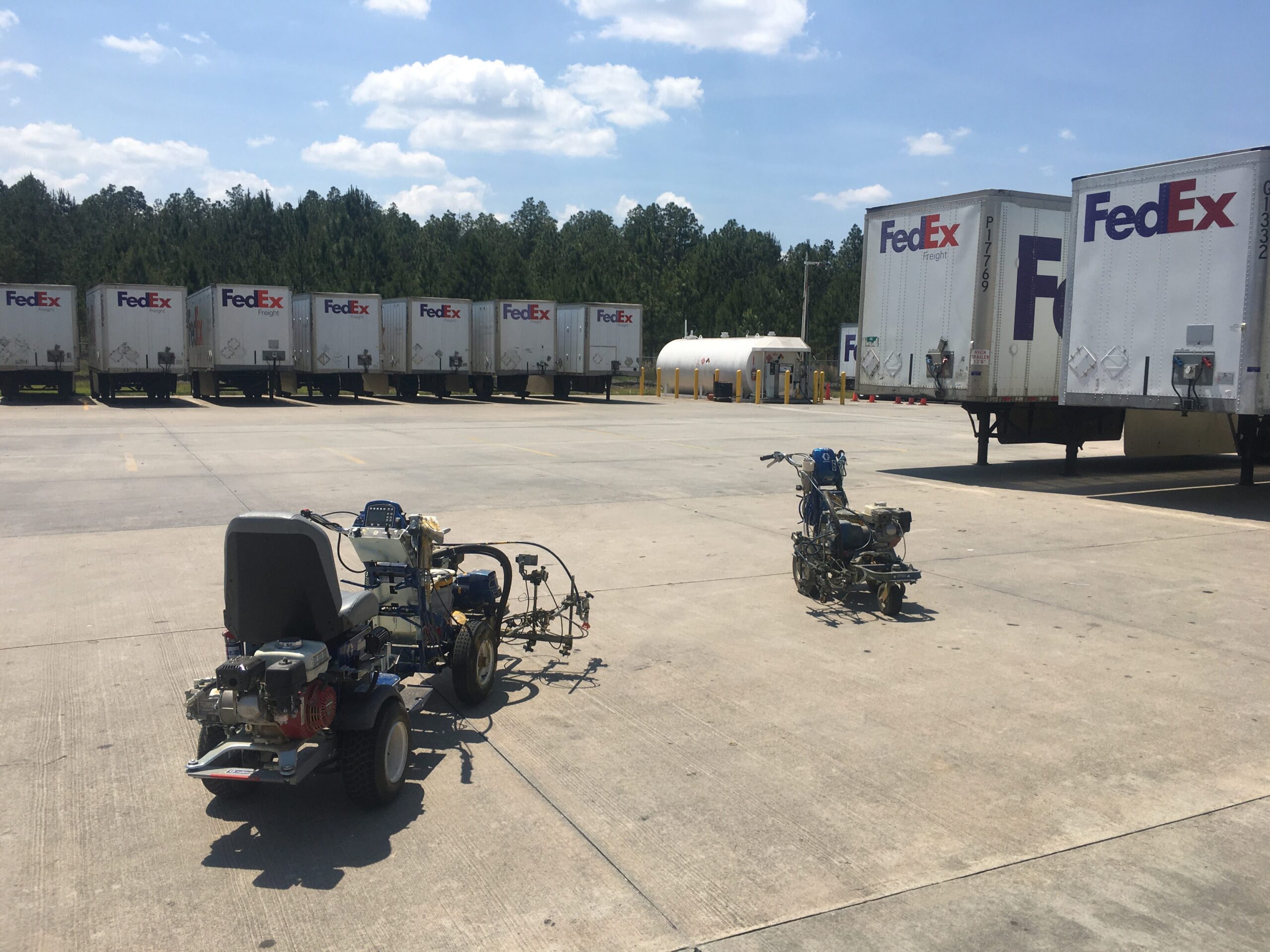 Line of FedEx semi trailers parked in a lot with a tree line in the background and equipment in the foreground.3eb; A row of white FedEx trailers lined up, with clear blue sky and scattered clouds above, and some machinery in the parking area.