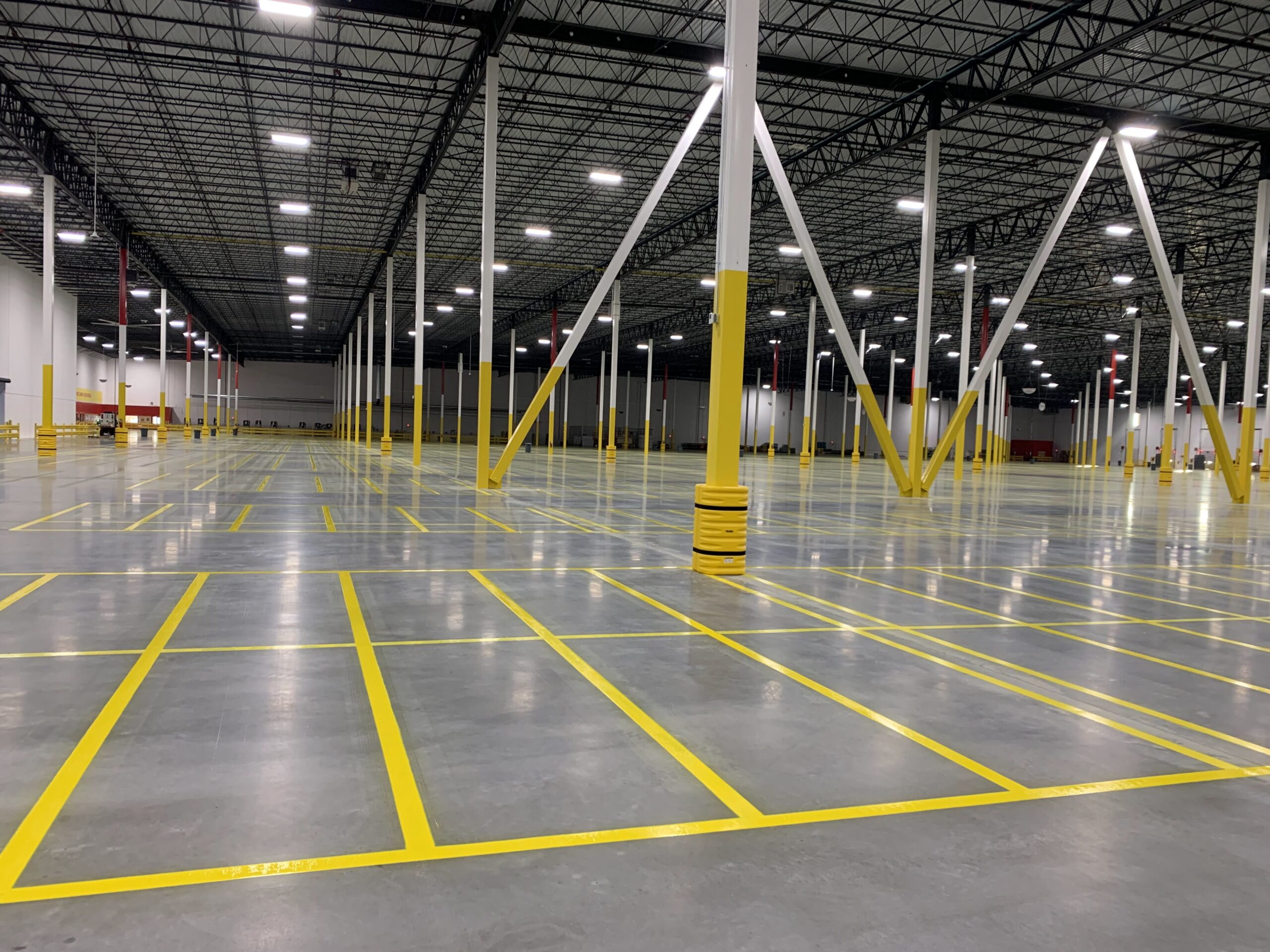 Wide empty warehouse interior with yellow safety markings and white support columns under a metal roof structure, well-lit scene