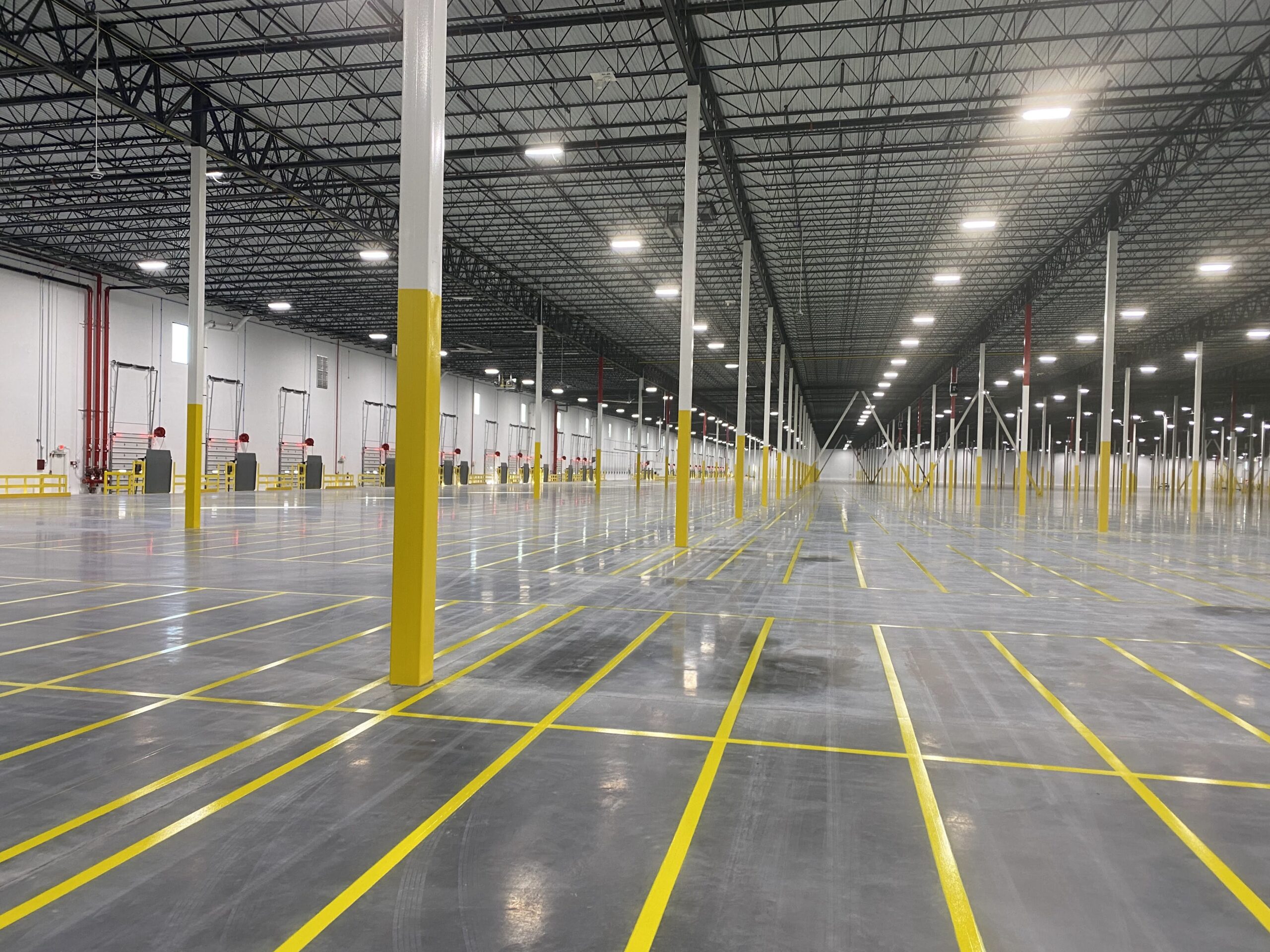 Empty warehouse interior with tall white columns painted yellow at the base and numerous yellow floor markings forming lanes or aisles under a metal-truss ceiling.