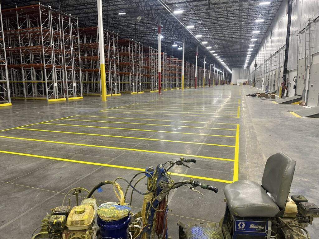Wide interior of a large warehouse with tall metal pallet racks along the left yellow safety posts and bright overhead lights bicycles in the foreground