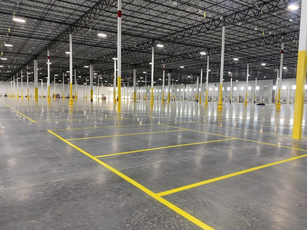 Wide view of a large empty warehouse with yellow floor markings and numerous white columns with yellow bases under a high metal ceiling of crisscrossing beams
