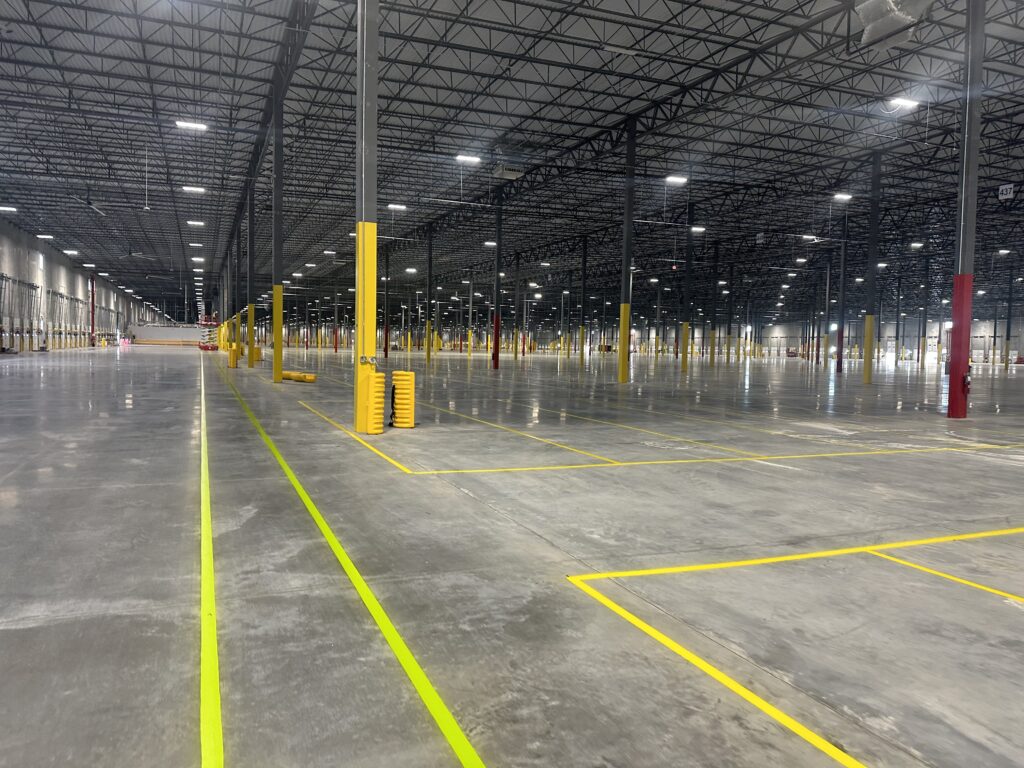 Empty warehouse interior with tall steel columns, overhead trusses, and bright yellow safety posts and floor markings.